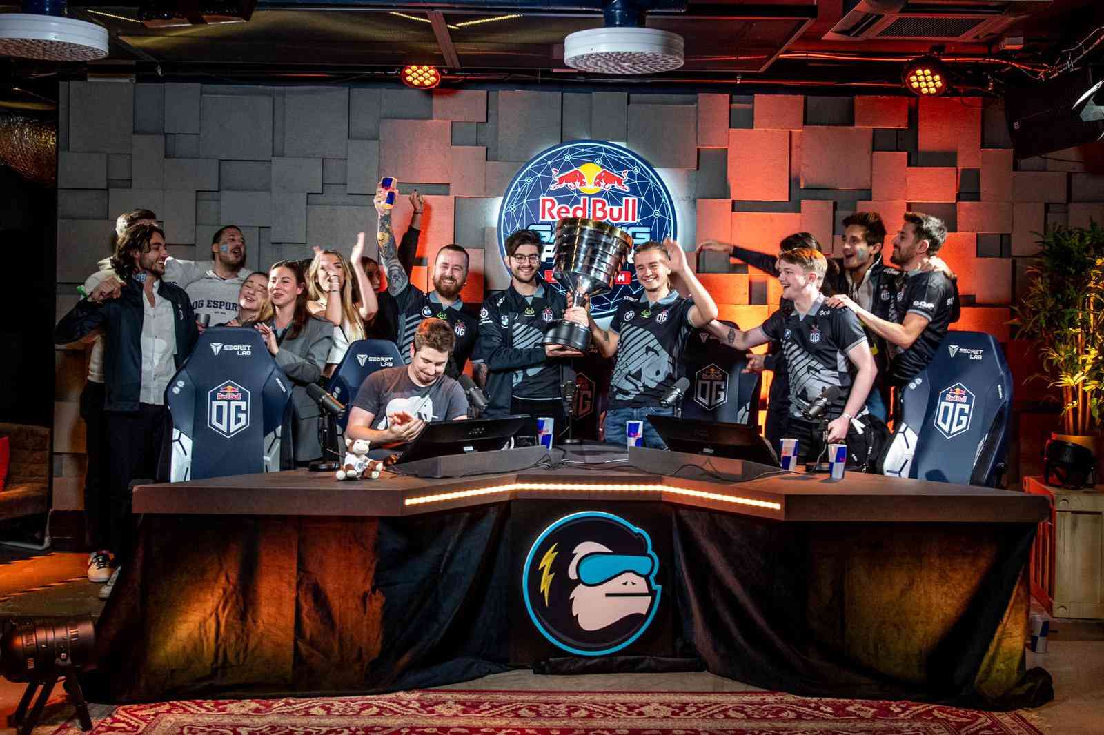 OG players and staff stand together around a desk on stage, cheering and holding memorabilia and the ESL trophy